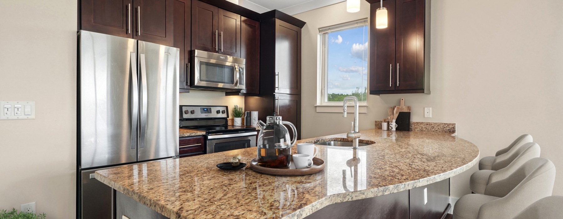 Wide banner view of a modern open-concept apartment interior at Matrix Hudson, showcasing a gourmet kitchen island with seating flowing into the living area in Hudson, MA.